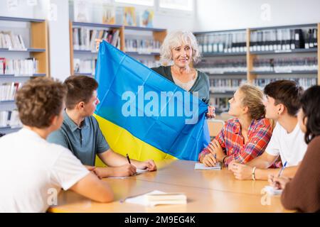 Interesting story about Ukraine while teaching high school students in school library. Teacher holds flag of Ukraine in her hands Stock Photo