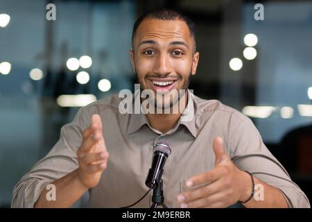 Successful hispanic man inside office looking at camera and talking on video twin inside office, businessman recording audio podcast using professional microphone, webcam view pov. Stock Photo