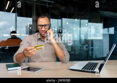 Dissatisfied businessman in the office with a bank credit card calls customer support, blocked card man can't make a purchase in an online store. Stock Photo