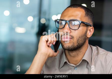 Serious and focused african american businessman close up in the middle of the office talking on the phone, a man in a casual shirt and glasses is listening to a client interlocutor. Stock Photo