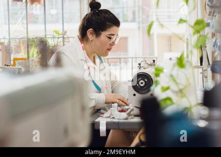 Caucasian young woman clothing designer concentrated sitting working with her sewing machine in her clothing workshop, with the door and window in the Stock Photo