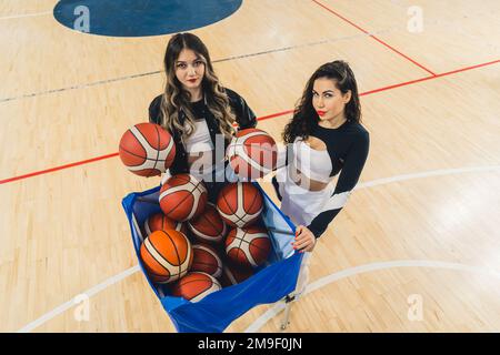 Top view of two female cheerleaders in cheerleader outfit posing with basketballs on basketball court. The rest of basketballs in blue basket. High quality photo Stock Photo