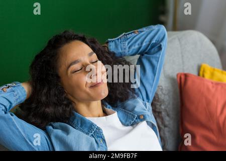 Beautiful hispanic woman at home close up with closed eyes relaxing sitting on couch, woman with hands behind head smiling in living room. Stock Photo