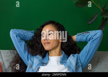 Beautiful hispanic woman daydreaming sitting on couch at home and looking out window, happy woman with hands behind head close up. Stock Photo