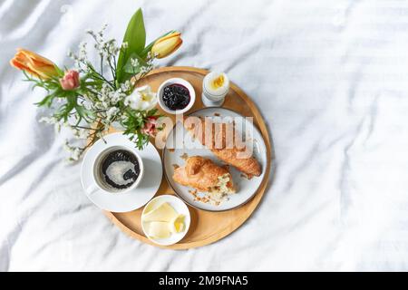 Lazy breakfast in bed, wooden tray with croissant, coffee, jam, egg, butter and flowers on gray green white bed linen, Sunday or holiday morning, copy Stock Photo