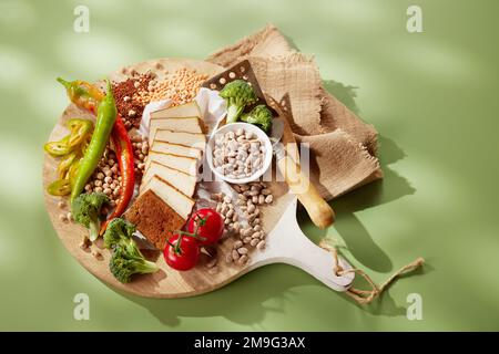 From above of appetizing sliced cheese with raw broccoli and cherry tomatoes served on round shaped wooden cutting board with scattered nuts and chili Stock Photo