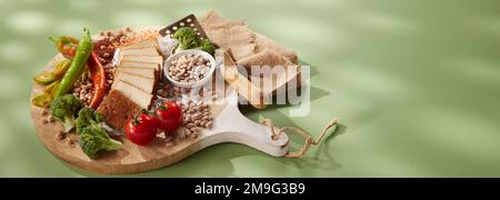 High angle of various delicious healthy raw vegetables seeds and nuts arranged on round wooden cutting board with sliced cheese and spatula on green t Stock Photo