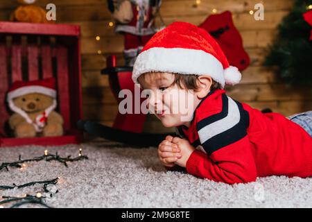 close-up of the face of cheerful blond Caucasian boy in a red cap lying on a wooden background at Christmas Stock Photo