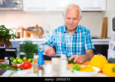 elderly man cuts vegetables for salad at the table in the kitchen Stock Photo