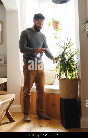 Smiling biracial bearded young man spraying water on potted plant by window at home Stock Photo