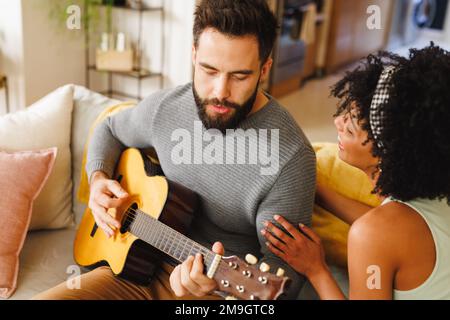 Biracial bearded young man singing and playing guitar while sitting with girlfriend on sofa Stock Photo