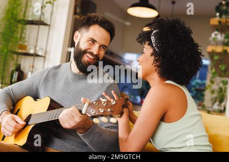 Biracial young woman with afro hair looking at boyfriend playing guitar while sitting on sofa Stock Photo