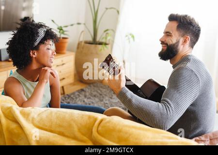 Happy bearded biracial young man playing guitar and singing while looking at girlfriend on sofa Stock Photo