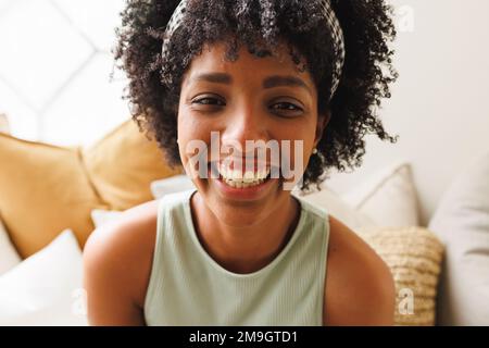 Close-up portrait of biracial smiling young woman with afro hair sitting on sofa at home, copy space Stock Photo