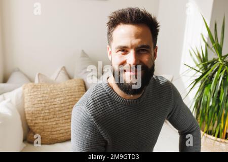 Portrait of smiling bearded biracial young man sitting on sofa in living room over wall, copy space Stock Photo