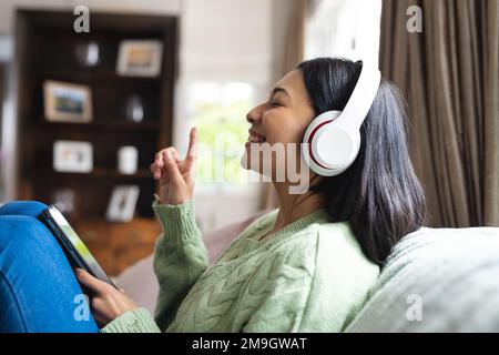 Image of happy biracial woman in headphones using smartphone sitting on sofa at home, copy space Stock Photo