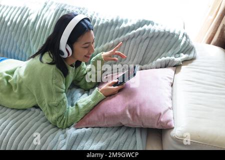 Image of happy biracial woman in headphones using smartphone lying on sofa at home, copy space Stock Photo