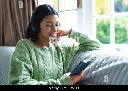 Image of happy biracial woman in headphones using smartphone sitting on sofa at home, copy space Stock Photo
