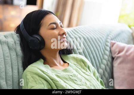 Image of happy biracial woman in headphones relaxing on sofa with eyes closed at home, copy space Stock Photo