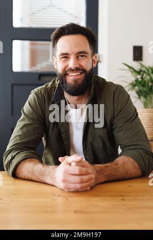 Portrait of biracial bearded young man wearing jacket with hands clasped sitting at table Stock Photo