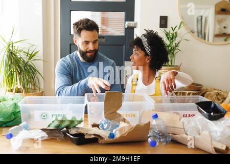 Biracial young couple discussing and sorting papers, plastics and glasses wastes in bins on table Stock Photo