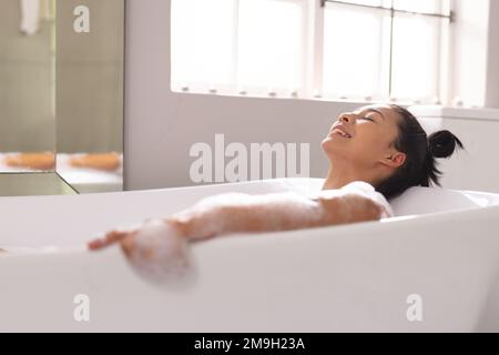 Image of smiling biracial woman in bathtub in bubble bath in bathroom Stock Photo