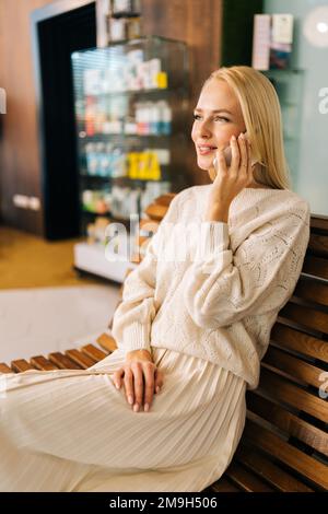 Vertical portrait of happy smiling young woman talking on mobile phone sitting on wooden bench. Beautiful woman having smartphone call indoor hotel. Stock Photo