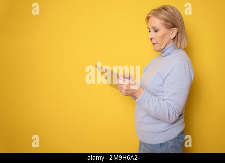 Portrait of smiling beautiful senior aged mature woman using tablet computer, isolated over yellow background. Copy space for text. Stock Photo