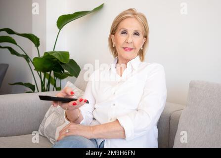 Cheerful senior woman enjoys evening with a TV shows, series. Smiling mature woman sits on the sofa with TV remote controller and switching channels Stock Photo