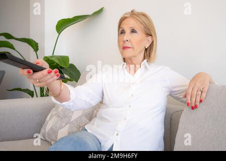 Cheerful senior woman enjoys evening with a TV shows, series. Smiling mature woman sits on the sofa with TV remote controller and switching channels Stock Photo