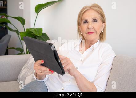 Smiling mature old woman holding using digital tablet relaxing sit on sofa reading e book in reader app, happy middle aged senior grandma browsing int Stock Photo
