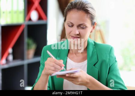 Confident and satisfied business woman writes notes in her notebook while sitting at office desk. Stock Photo
