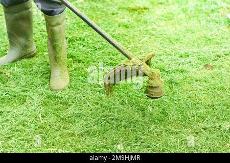 Man or worker mowing the green grass. Gardener mows weeds with electric or petrol lawn trimmer in city park or backyard. Gardening care tools and equi Stock Photo