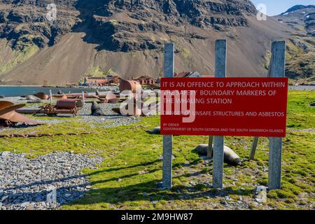 Asbestos warning sign on the beach in Stromness Bay on South Georgia Island, sub-Antarctica with the abandoned whaling station in the background. Stock Photo