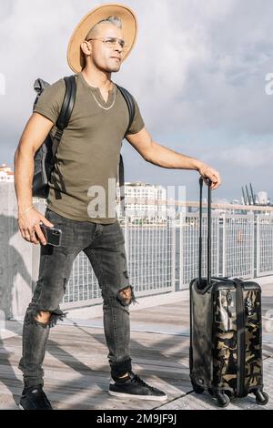 Portrait of a traveller man holding a smartphone and a carrying suitcase. traveller man wearing a hat, a t shirt and a ripped jeans, Hipster style. Stock Photo