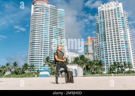 Hipster man with luggage bag on wheels standing on the south beach in miami. Sunny day, Travel concept Stock Photo