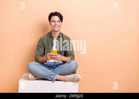 Full size body cadre of young student sitting platform smartphone proposition eshopping cheap clothes app offer isolated on beige color background Stock Photo