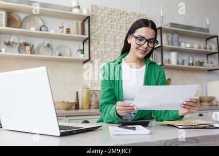 Young smiling woman designer, engineer, architect works from home online. Sitting in the kitchen at the laptop. Holds documents, plans, drawings in his hands. Stock Photo