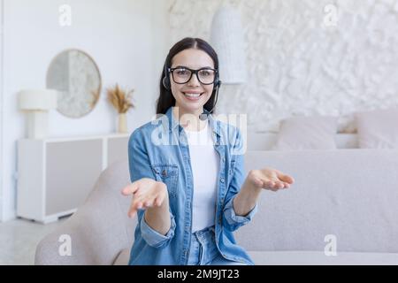 Young beautiful female blogger sitting on sofa at home in headset and chatting online on camera. He talks, gestures with his hands, smiles. Stock Photo