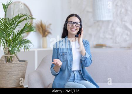 Distance Learning. Portrait of a young female teacher in glasses sitting at home on the sofa in a headset, talking and explaining to the camera. Works, teaches from home online. Stock Photo