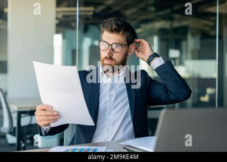 Middle aged male manager with documents checking marketing report, having problems, sitting at desk in office Stock Photo