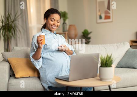 Positive young pregnant black woman video calling to doctor and showing medical pills bottle to laptop, sitting on couch Stock Photo