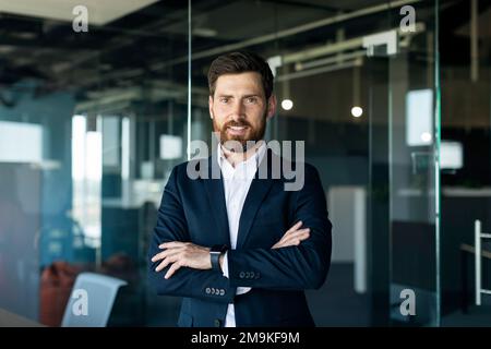 Portrait of confident smiling middle aged businessman in formal wear posing with crossed hands in office interior Stock Photo