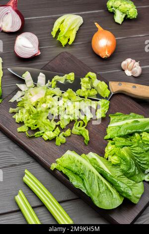 Chopped lettuce leaves and knife on cutting board. Onion, garlic and celery stalks on table. Dark wooden background. Stock Photo