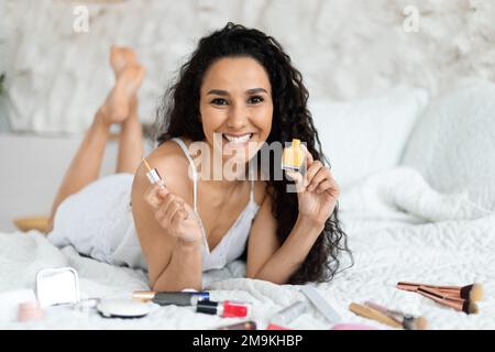Cheerful middle eastern millennial curly female in domestic clothes lies on bed with cosmetics and nail polishes Stock Photo