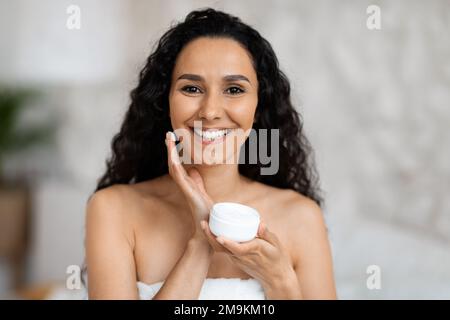 Portrait of cheerful middle eastern millennial curly lady in towel holding jar, applies cream on face Stock Photo