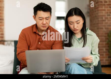 Upset korean spouses checking bills, reading documents while sitting on couch and using laptop. Family budget concept Stock Photo