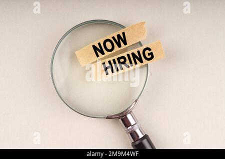 On the magnifying glass are paper strips with the inscription - Now hiring. Stock Photo