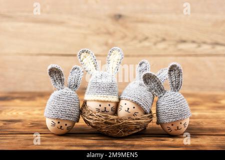 Four Easter eggs with funny faces and gray crocheted hats with bunny ears in a decorative nest of sisal on a wooden background. Happy Easter concept. Stock Photo