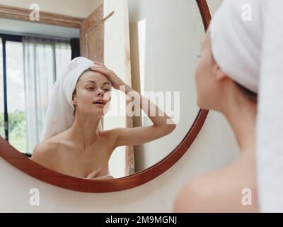 A beauty woman stands in front of a mirror after a shower in a towel on her head looks at her reflection and does a facial massage applies a day cream Stock Photo